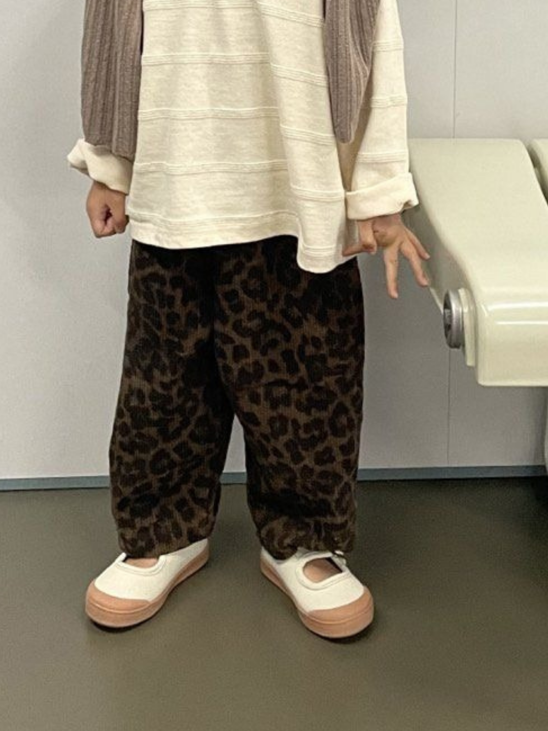 Dark | A child indoors next to a light chair showcases a stylish Korean brand look, wearing LEOPARD CORDUROY PANTS paired with a cream long-sleeve shirt and white shoes.
