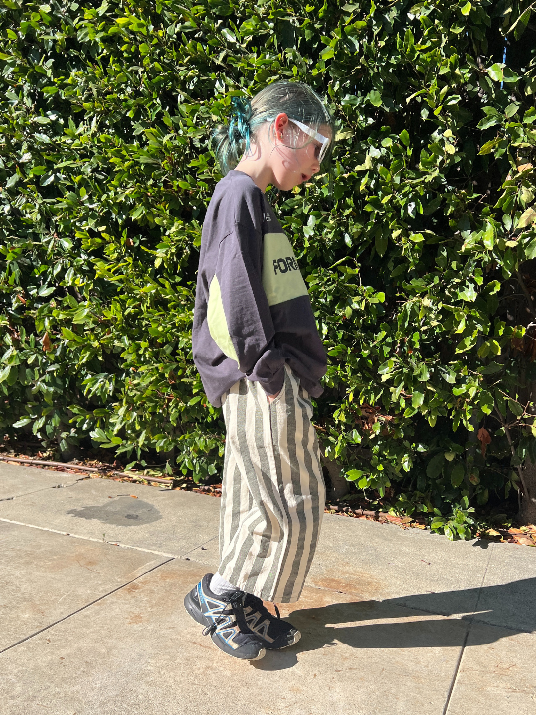 Person wearing a dark sweater with a logo and striped pants standing against a green hedge.