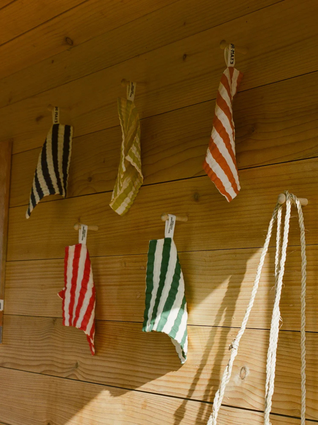 Five ORGANIC BABY WASH CLOTHS in various colors, made from soft muslin cotton, hang from hooks on a wooden wall, with two white ropes to the right.