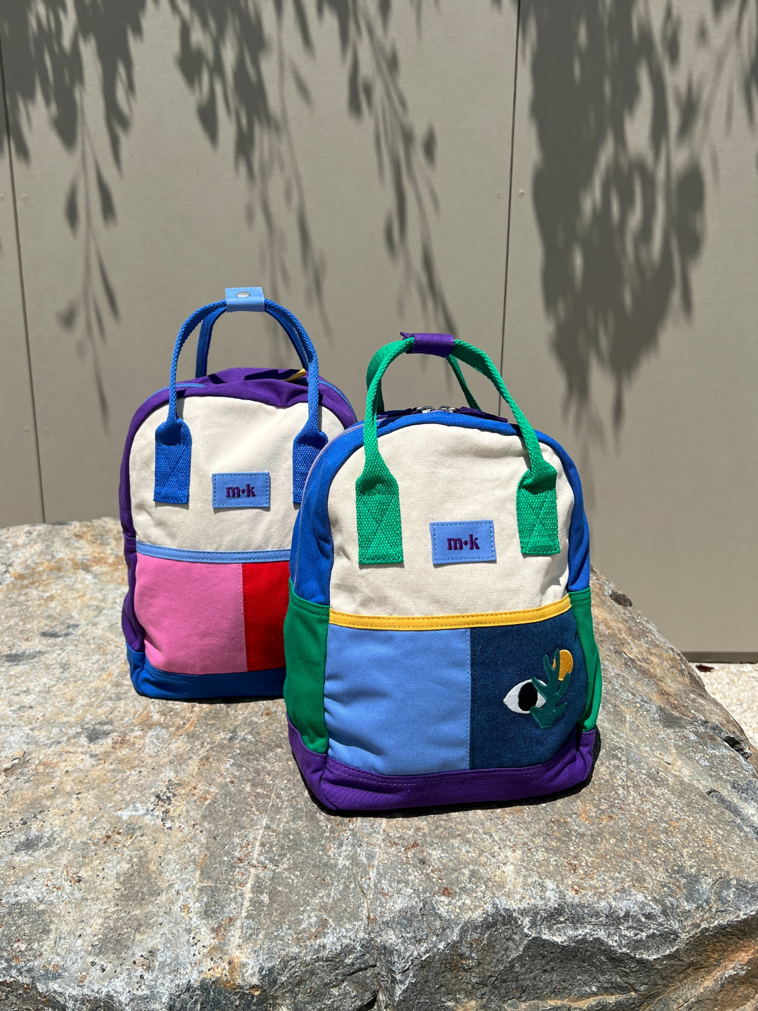 Two colorblock backpacks on a rock: one with blue handles and base, purple sides and one pink, one red patch under a cream top; one with green handles and sides, purple base, two blue patches under a cream top, one with images of an eye, a sun and a plant