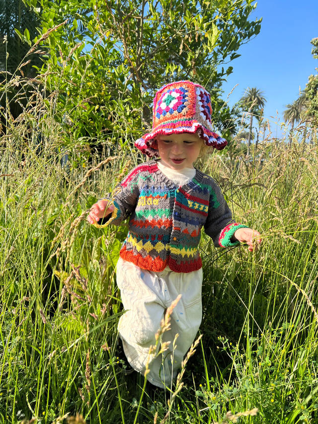 A young child stands in tall grass, wearing the HAND-CROCHETED BUCKET HAT - 1-3Y with a colorful sweater and white skirt, surrounded by greenery and blue sky.