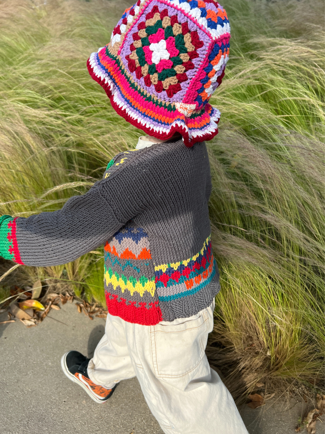 A child wearing the HAND-CROCHETED BUCKET HAT - 1-3Y and a patterned sweater walks outdoors beside tall green grass.