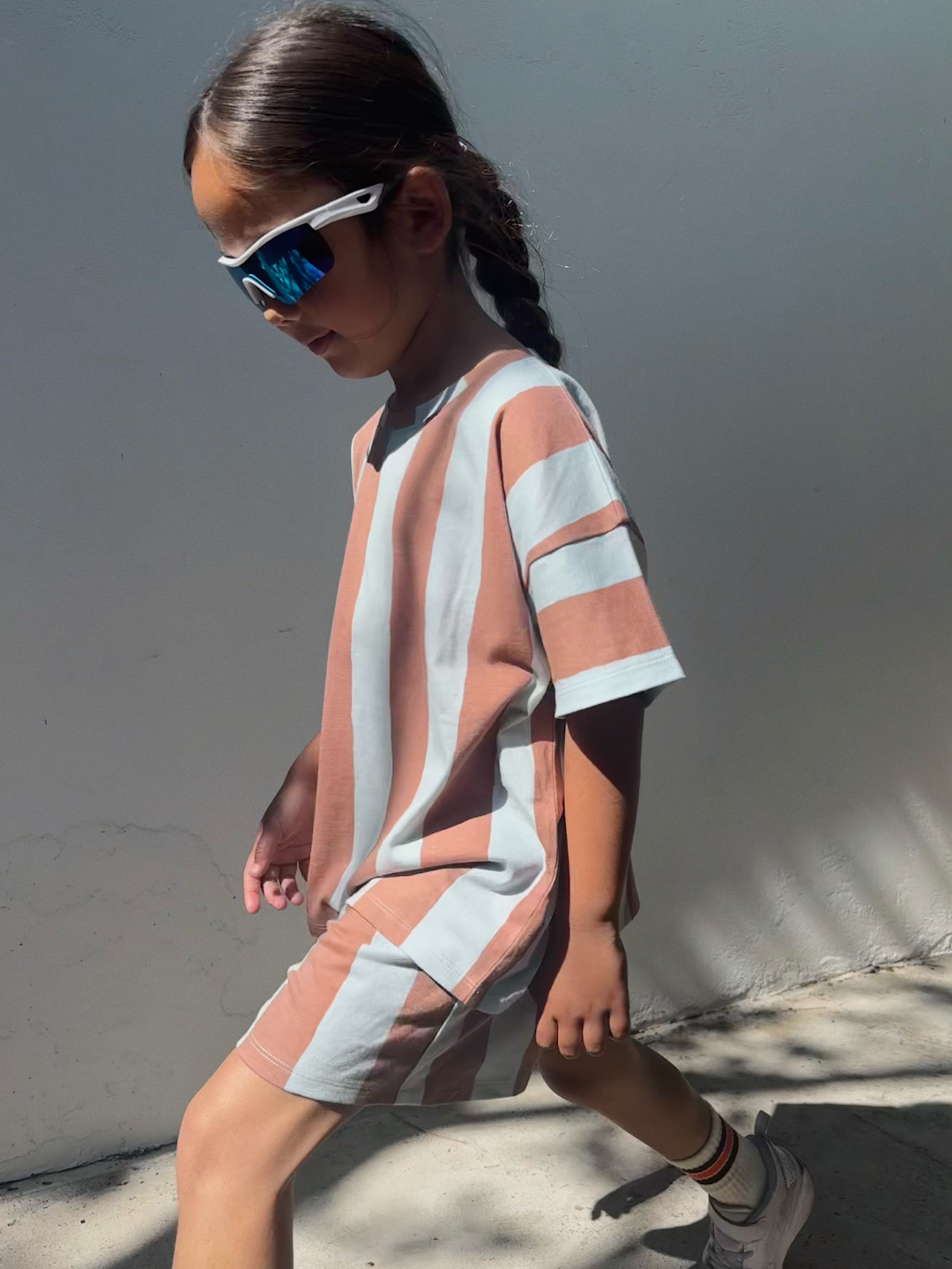 A child in the UMBRELLA STRIPE SHORT SET, sunglasses, and sneakers walks against a plain wall in sunlight, wearing blue sunglasses.