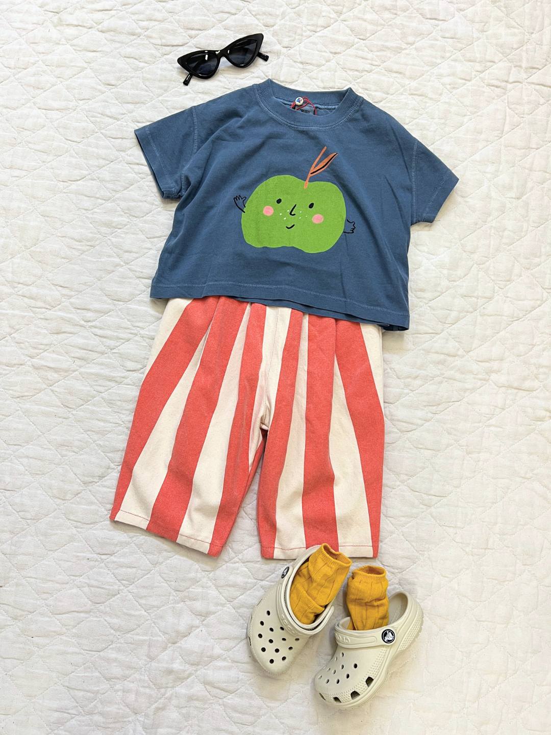Faded Red | Children's outfit laid flat: blue t-shirt with a green apple design, BIG STRIPE PANTS in faded red and summery colors, white sandals, yellow socks and black sunglasses on a white quilted background.