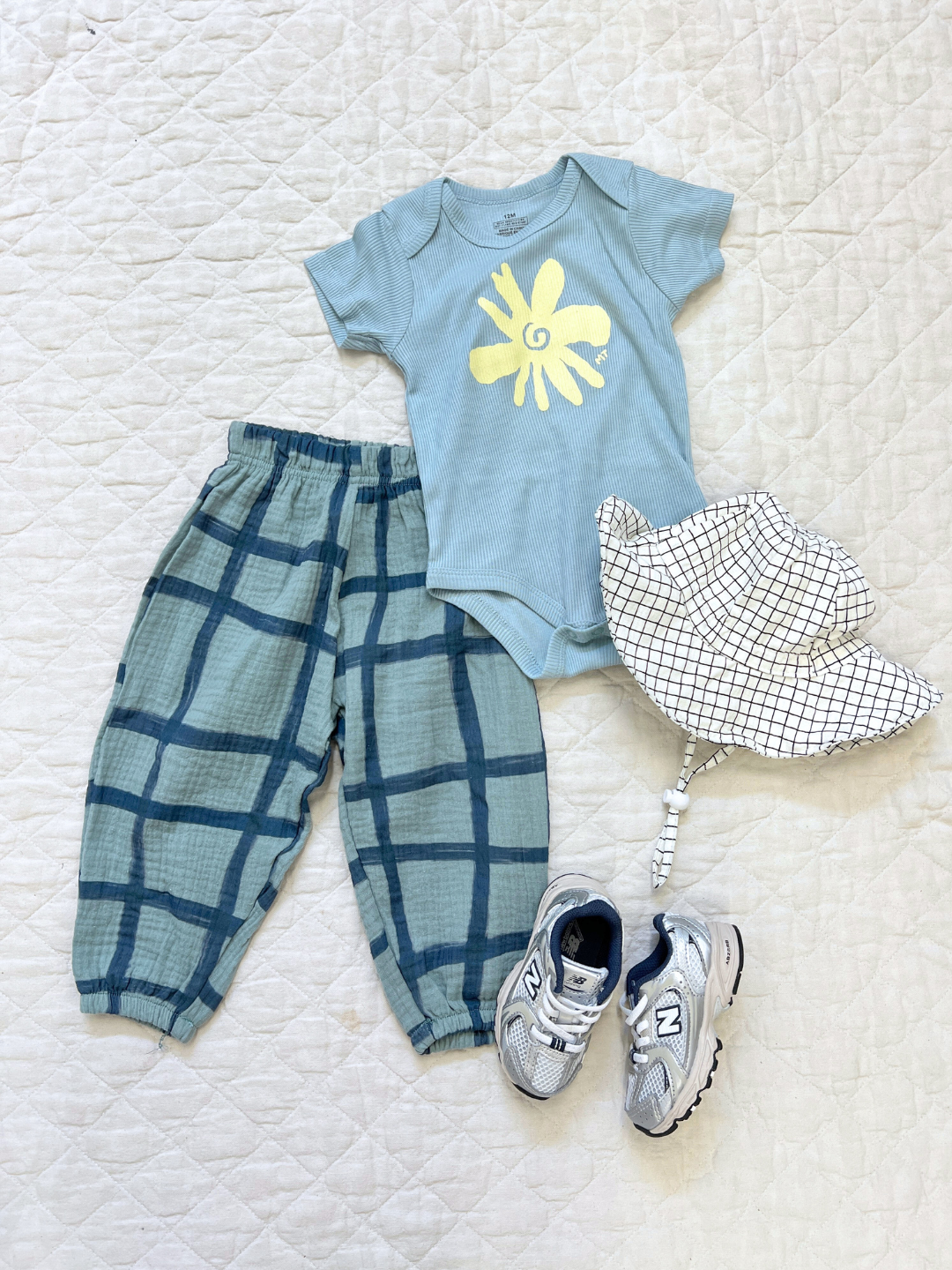 White | A flat outfit with the baby off the grid hat in white, paired with check pants and a flower onesie and grey sneakers, laid on a white cloth.