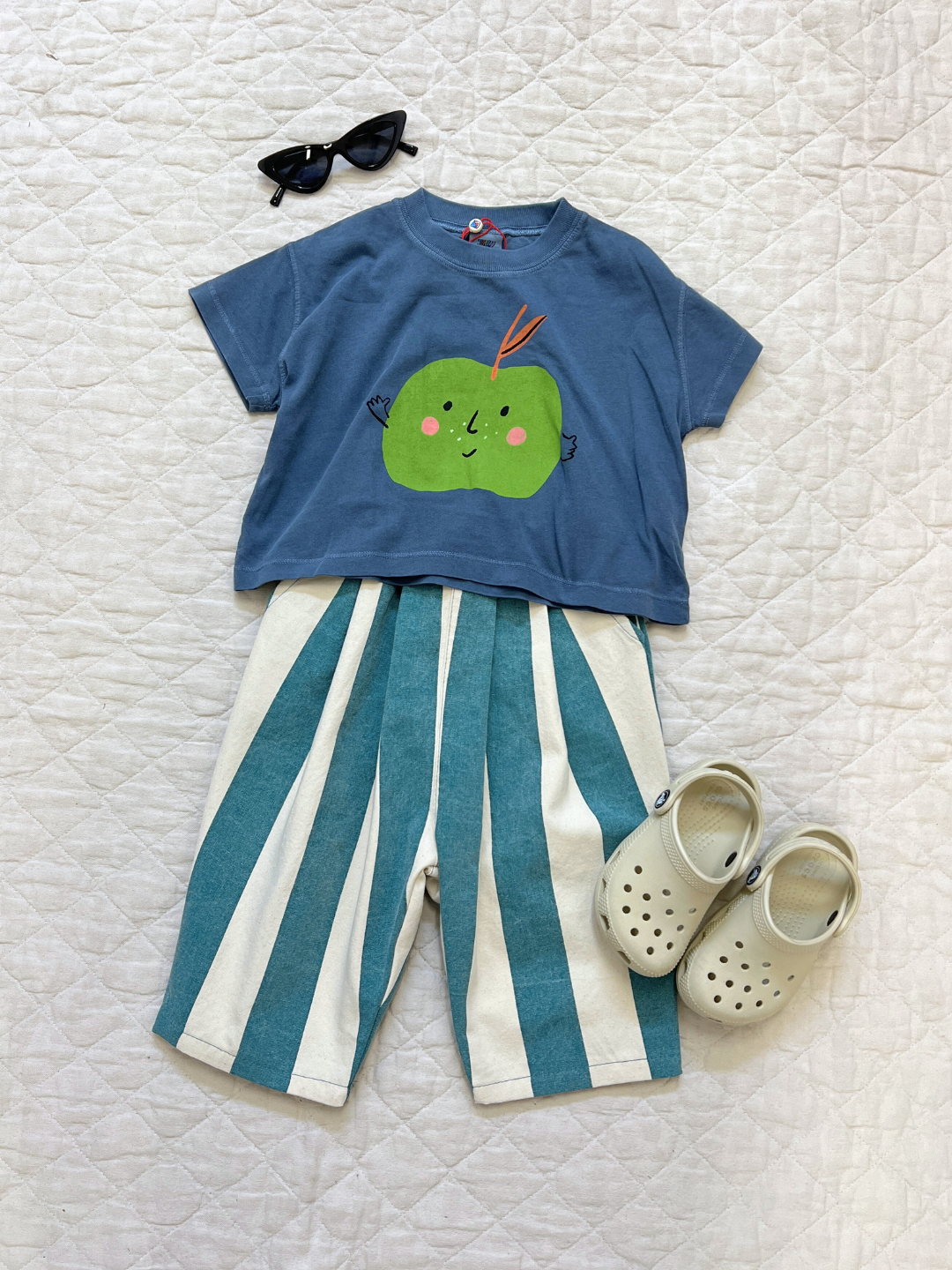 Teal | Children's outfit laid flat: blue t-shirt with a green apple design, BIG STRIPE PANTS in teal and summery colors, white sandals, and black sunglasses on a white quilted background.