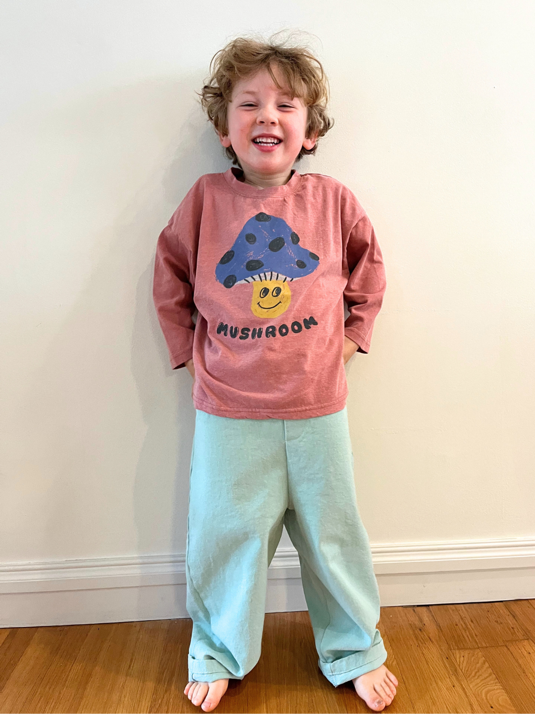 Mint | Child wearing the Billie Pants in mint, made from soft cotton twill with an elastic waistband and a yellow front button, with a red t-shirt with a mushroom character, standing against a white wall.