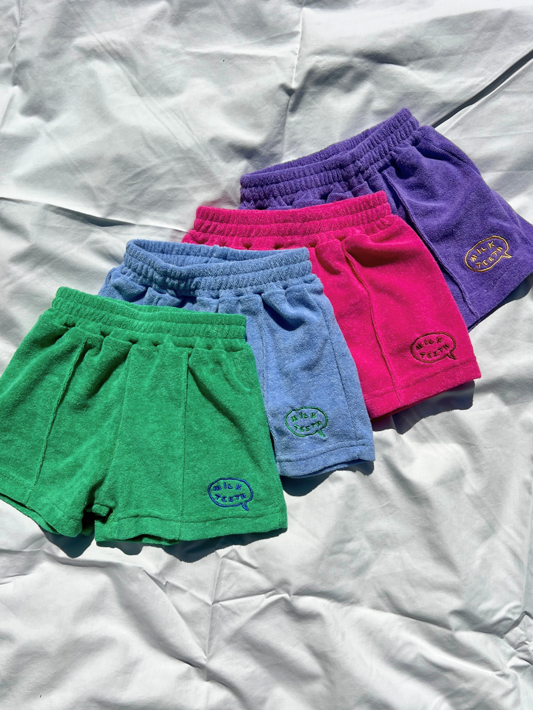 Four pairs of EVERYDAY TERRY SHORTS for kids, one green, one light blue, one pink and one purple, rest on a white cloth. They have elastic waists, cotton terry fabric, and small logos on the left leg.