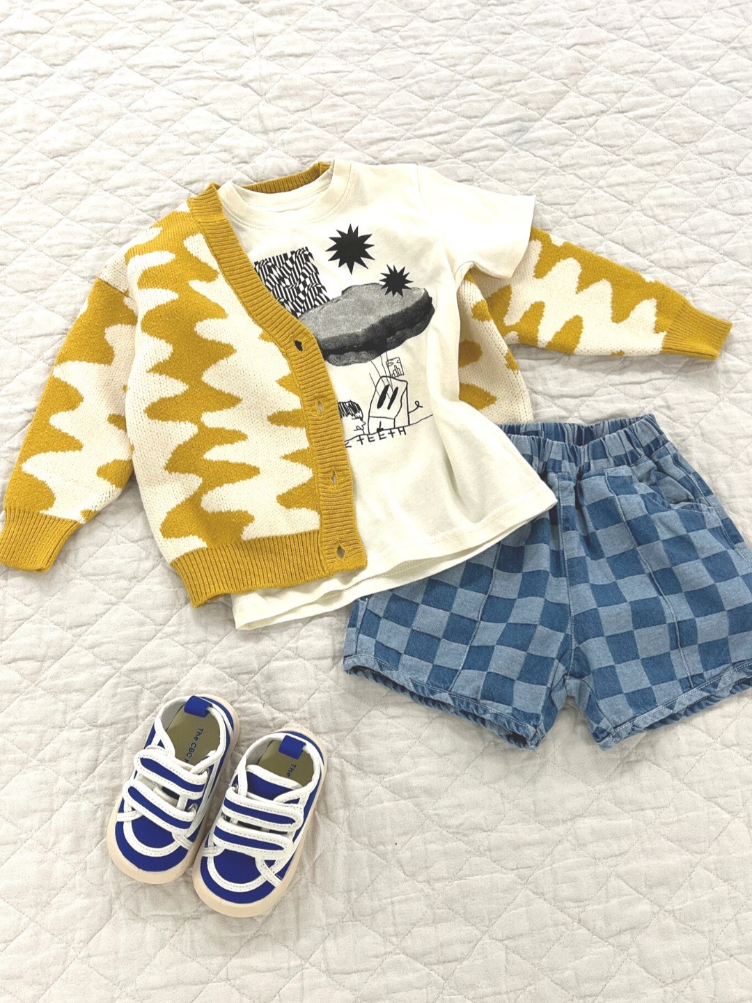 A child's outfit laid on a quilt showing a Wavelength Cardigan in marigold over a Sandwich Tee and a pair of kids' checkerboard shorts in two shades of blue, with a pair of Bounce House Sneakers in blue with white trim