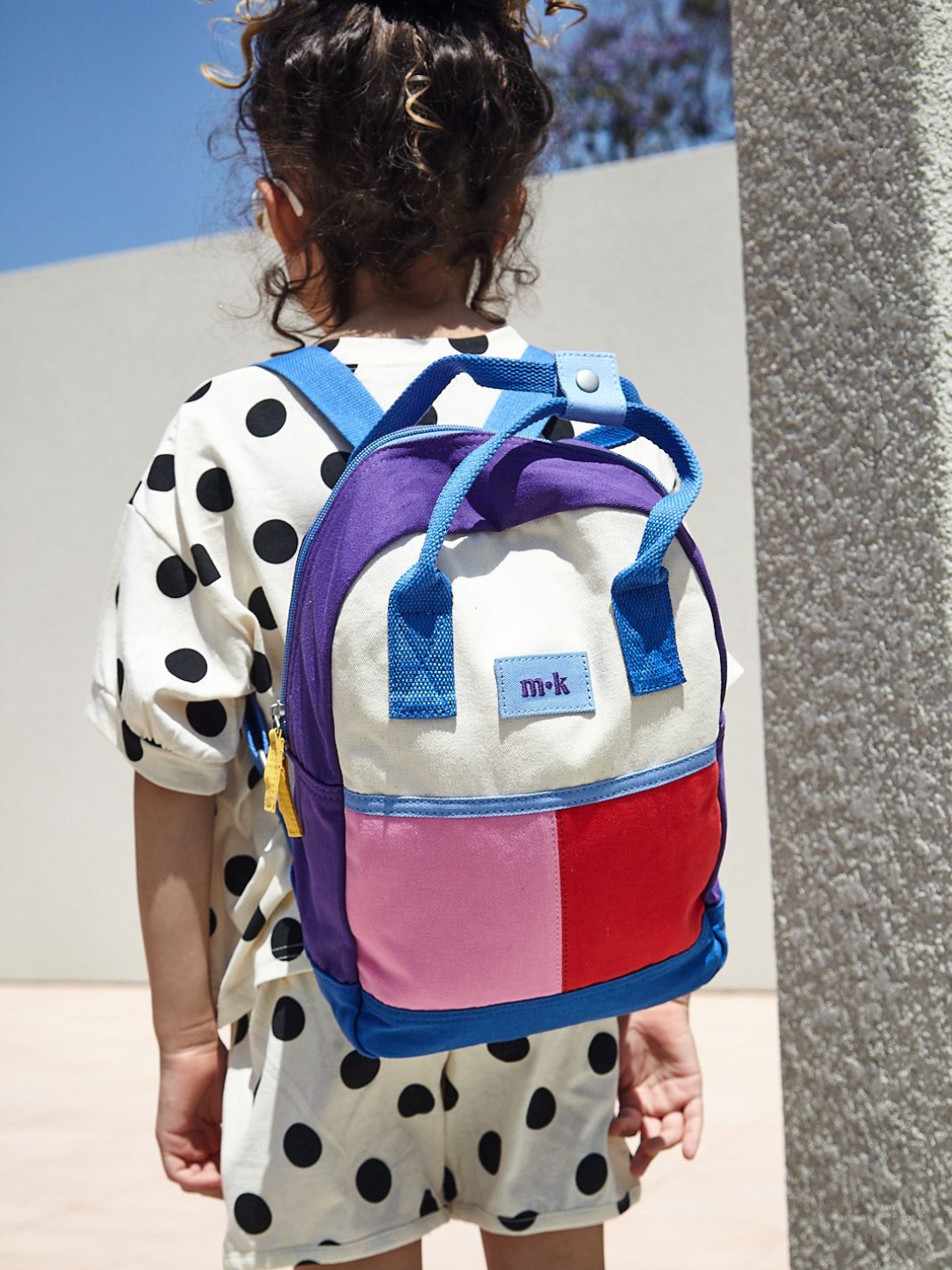 A child wearing a colorblock backpack with bright blue straps, handles and base, purple sides, and one pink, one red patch under a cream top