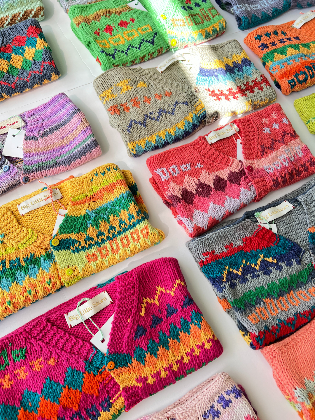 Rows of colorful, patterned HAND-KNITTED COTTON CARDIGAN - 2-3Y, made from soft cotton yarn, are neatly folded and labeled on a white surface.