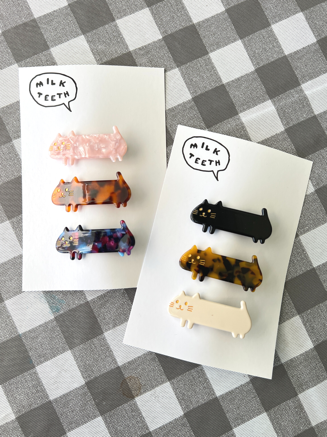 Two Milk Teeth presentation cards, each with three kids' barrettes in the shape of elongated cats; one pink, ginger and purple, one black, tortoiseshell and white