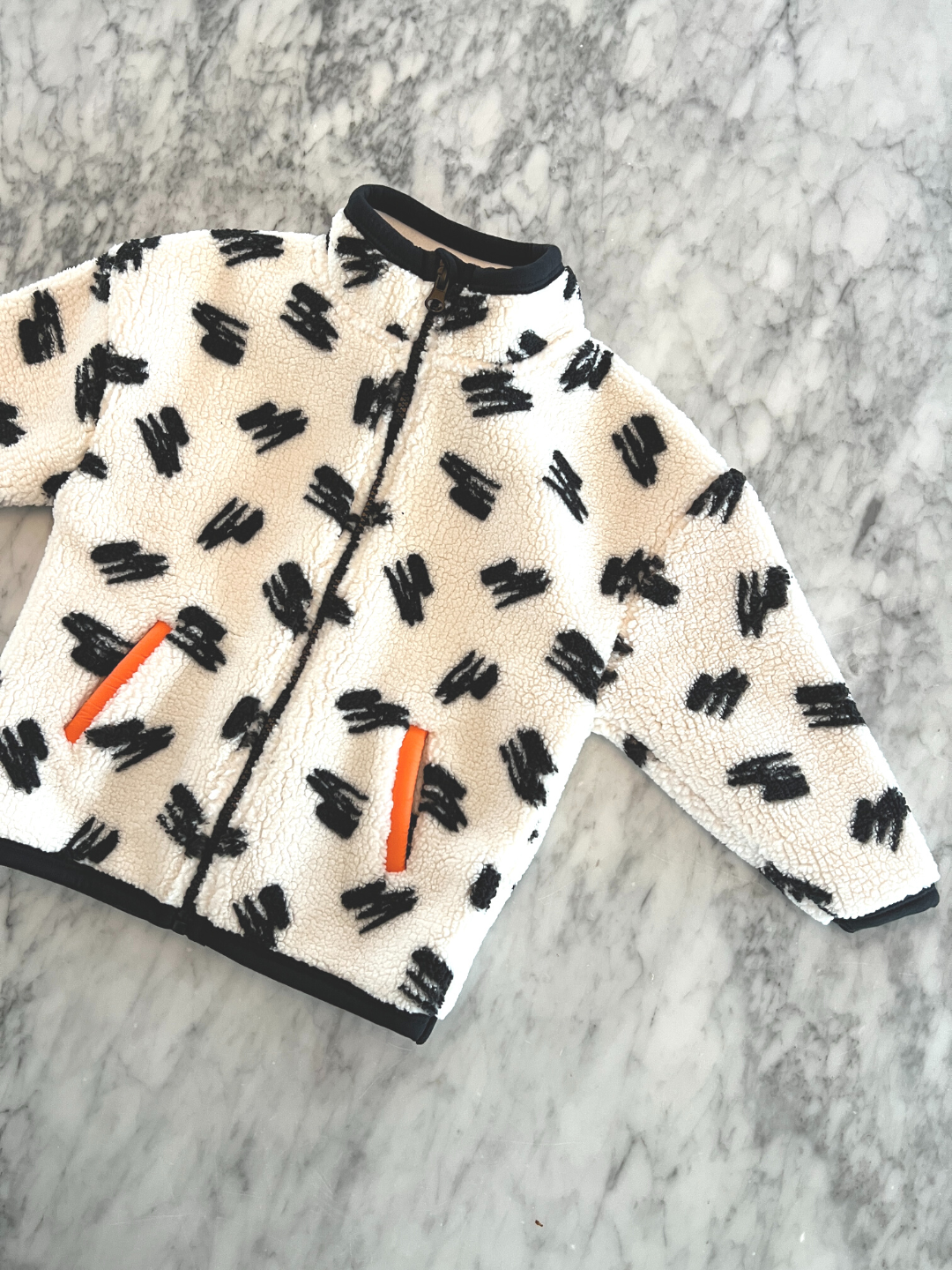 A kids' jacket laid on a table, patterned with black scribbles on a cream background, black binding on cuffs and hem, and orange binding on the pockets