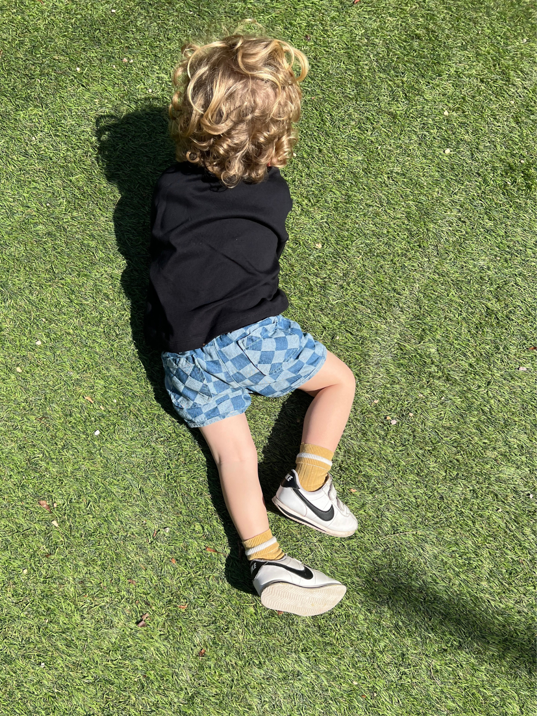 A toddler lying on grass wearing a pair of kids' checkerboard shorts in two shades of blue, back view