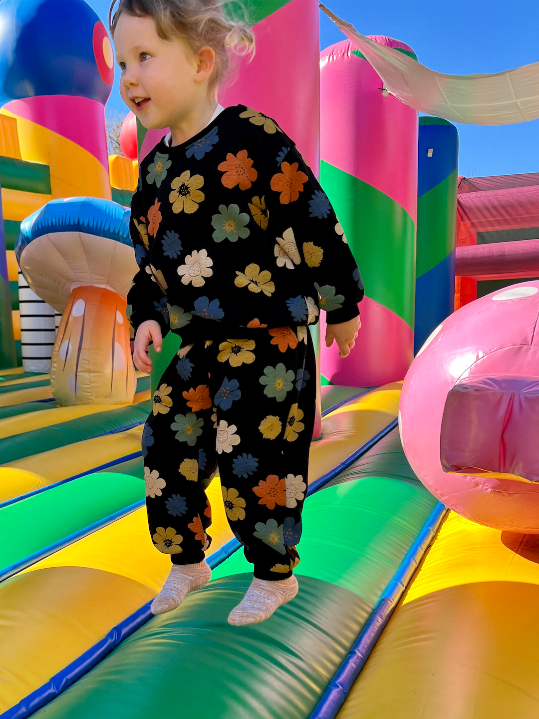A child on a bouncy castle wearing a kids' sweatshirt and pants set in black, printed with yellow, orange, green, blue and white flowers