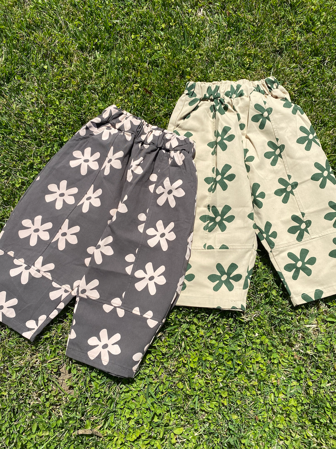 Black | Two pairs of kids' pants laid on grass: one in gray with cream flowers, one in cream with green flowers