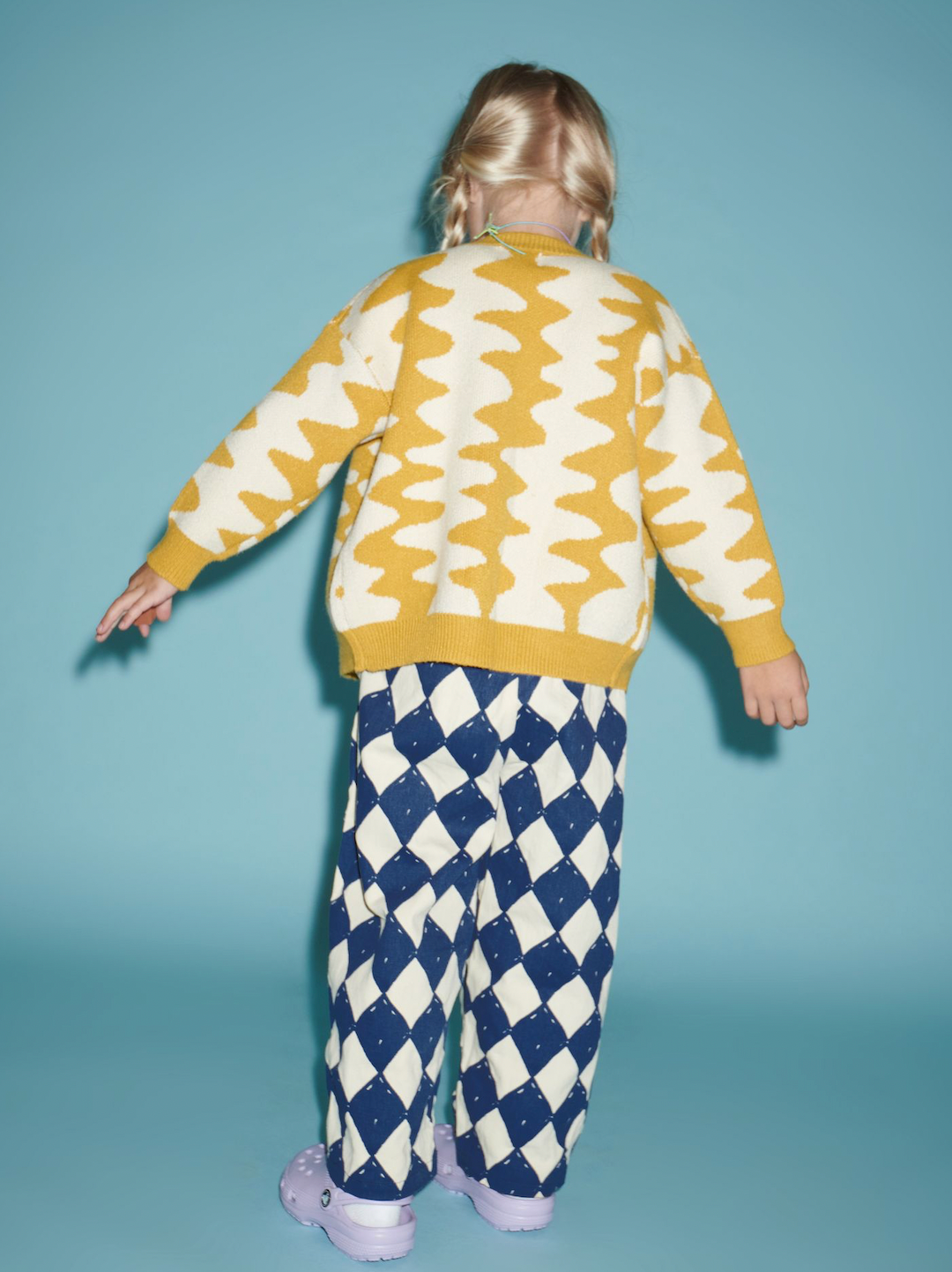 Marigold | A girl standing facing backwards, wearing Marigold Wavelength Cardigan with diamond pants and purple crocs.