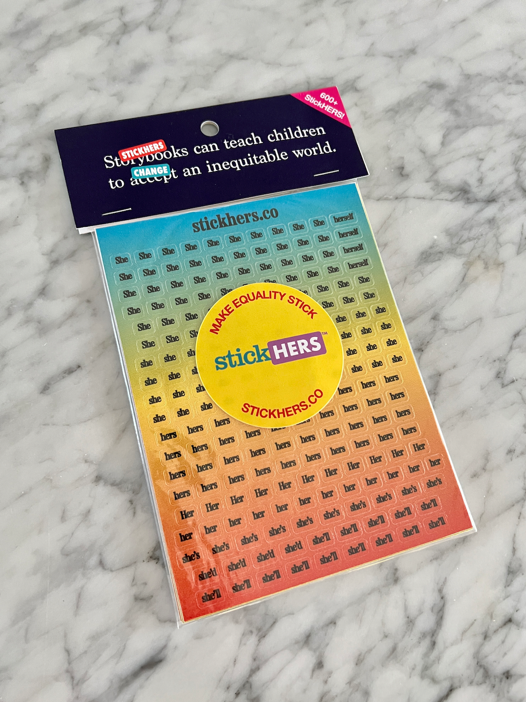 A pack of StickHERS colorful gender equity stickers displayed on a marble surface. The pack includes various pronouns, including non-gendered pronouns in different fonts and sizes. The packaging reads "Make Equality Stick" and "StickHERS.co" with a colorful gradient background.