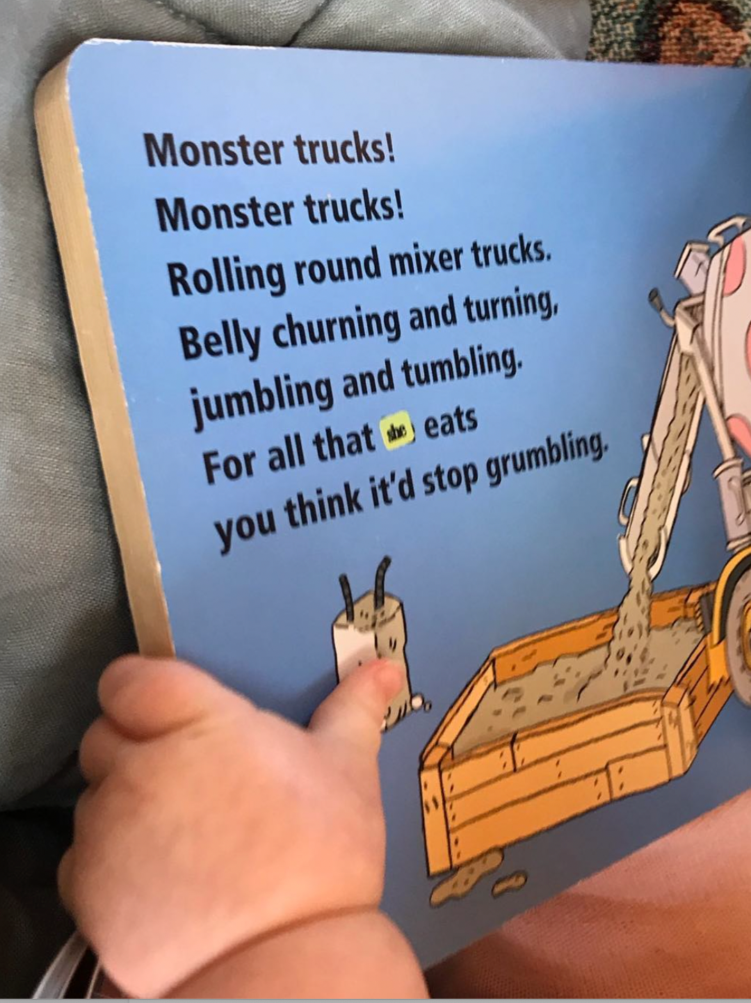 A close-up of a baby's hand holding a book page with an illustration of a monster truck pouring cement. The text on the page reads: "Monster trucks! Monster trucks! Rolling round mixer trucks. Belly churning and turning, jumbling and tumbling. For all that conc eats you think it'd stop grumbling." The page is adorned with StickHERS stickers, promoting gender equity.