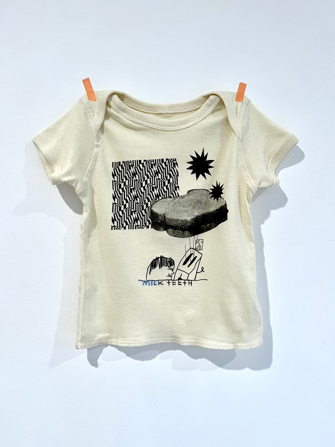 A baby tee shirt with lap shoulders, featuring patterns of black and white zigzags, stars, a PBJ sandwich, a toaster and the words Milk teeth