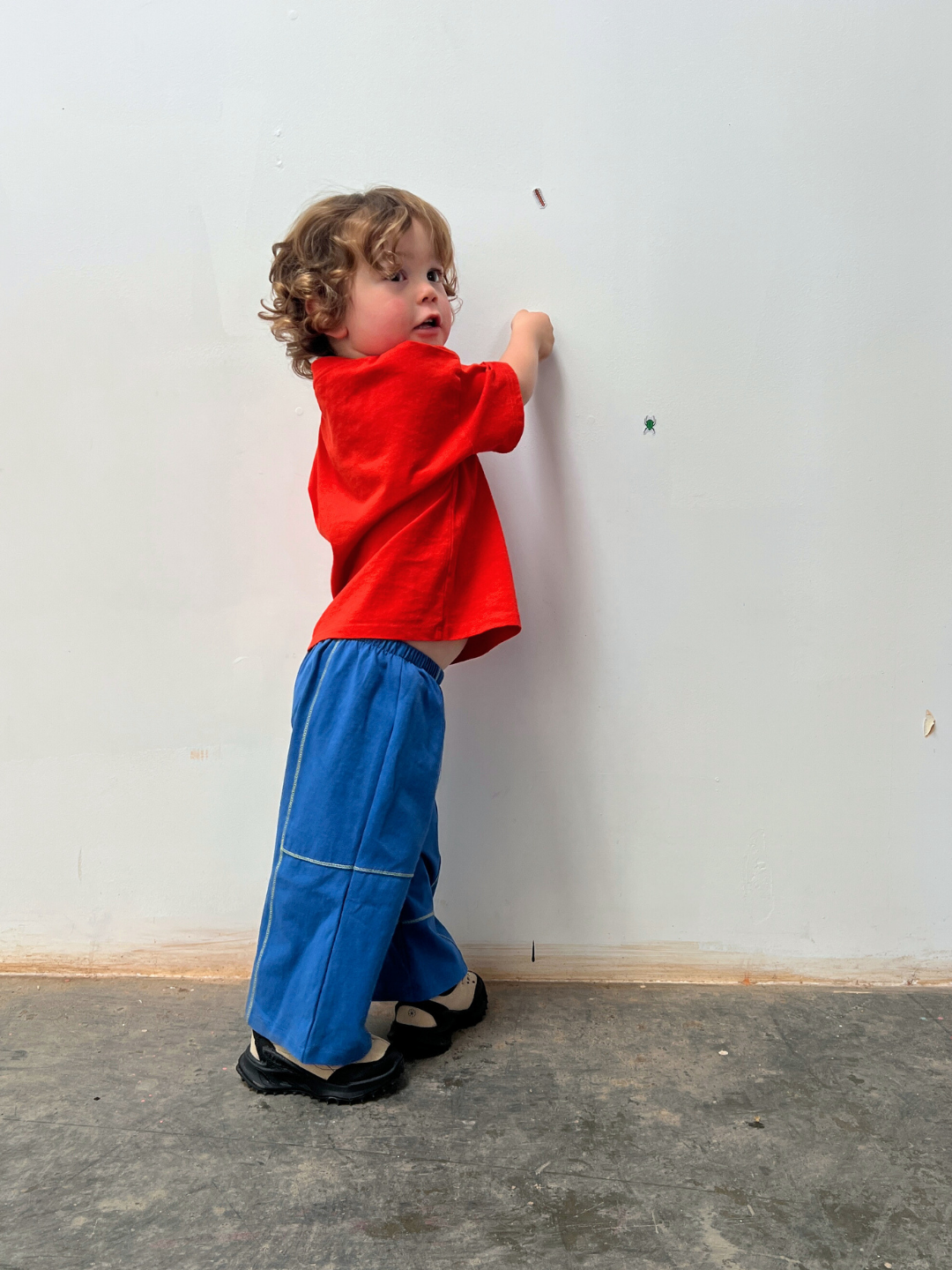 Blue | Child in a red shirt and blue pants standing against a white wall.