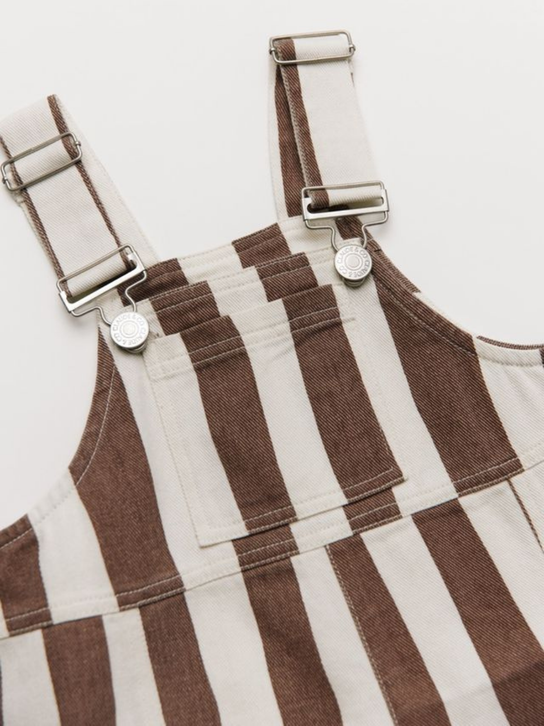 The BLYTHE CHOC STRIPE DUNGAREE features brown and white vertical stripes, denim fabric, and metal buckles and buttons, displayed flat on a white background.