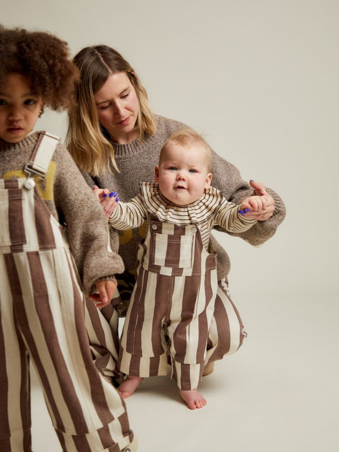 An adult woman supports a barefoot baby, both dressed in BLYTHE CHOC STRIPE DUNGAREE. Another child wearing the same dungaree stands to the left.