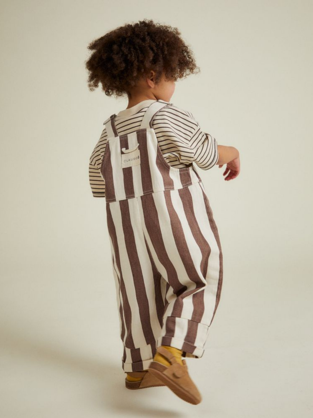 A young child with curly hair wears the BLYTHE CHOC STRIPE DUNGAREE, a striped shirt, yellow socks, and brown shoes, standing against a plain light background.