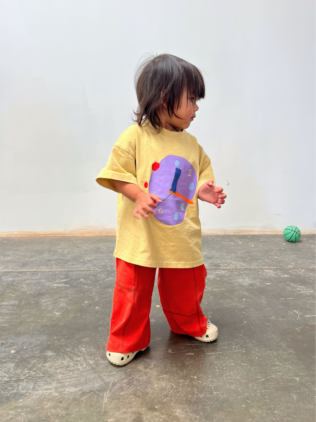 Child wearing a yellow shirt with a colorful design and red pants standing on a gray floor.