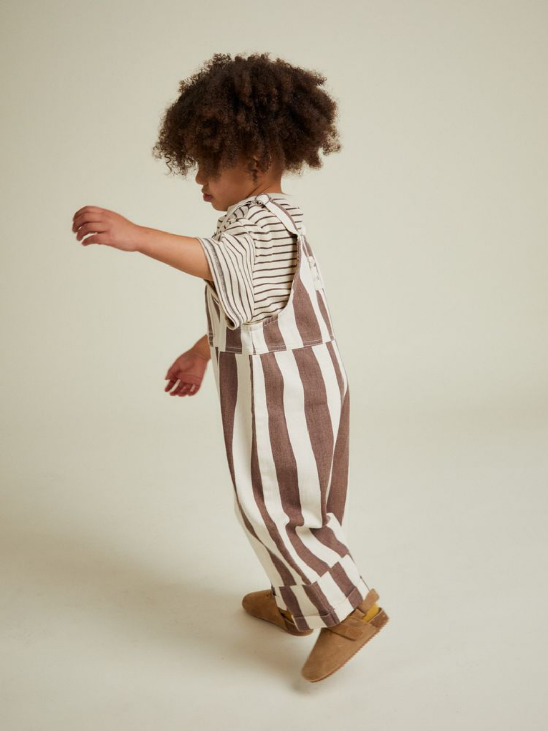 A young child with curly hair wears the BLYTHE CHOC STRIPE DUNGAREE and a striped short-sleeve shirt with brown shoes, walking against a plain background.