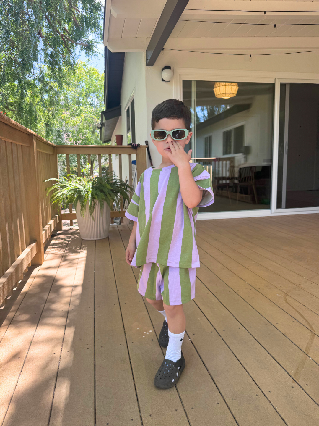 Green/Purple | A young child in sunglasses makes a peace sign while wearing the UMBRELLA STRIPE SHORT SET, a green-and-lavender striped outfit made from organic cotton, standing on a wooden deck.