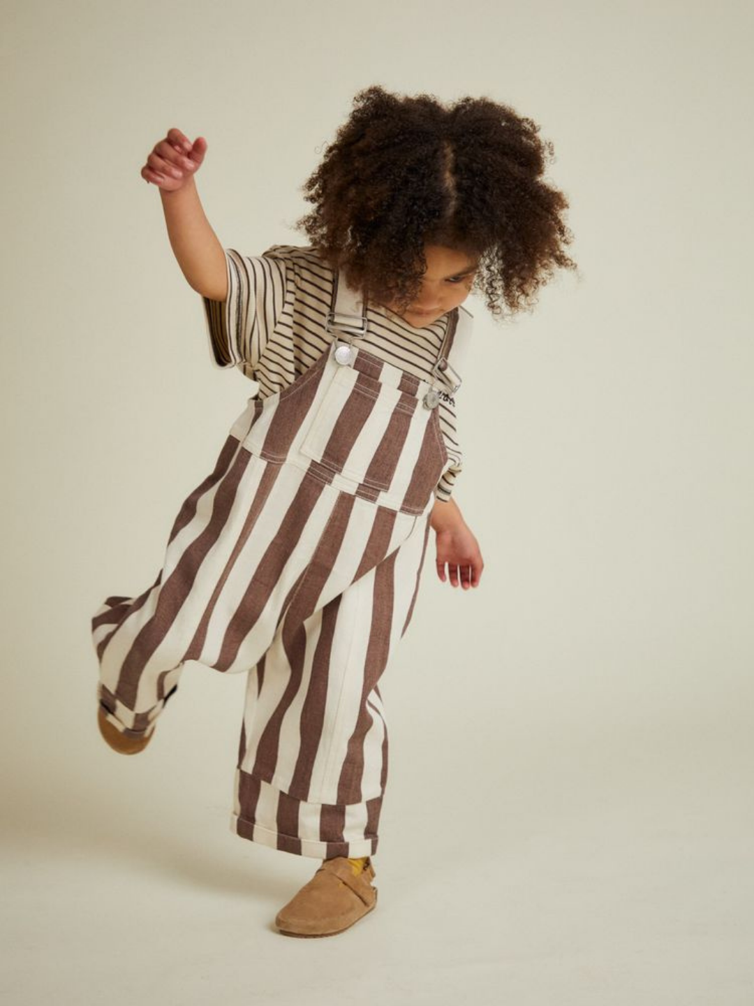 A young child with curly hair models the BLYTHE CHOC STRIPE DUNGAREE, standing on one foot against a plain background.