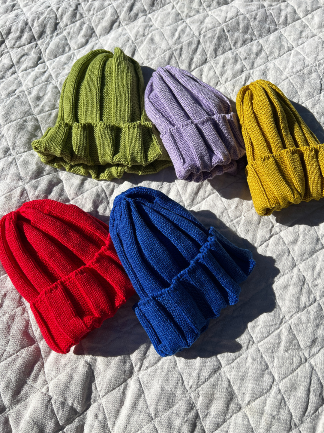 A selection of kids' knitted beanies laid on a quilt; green, lilac, mustard, red and blue