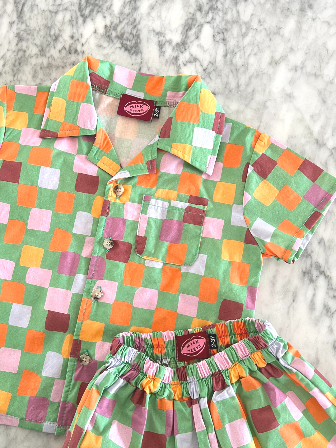 Green | Close-up of a kids' shirt and shorts set in a pattern of purple, pink, gold and orange squares on a green background, showing collar and pocket