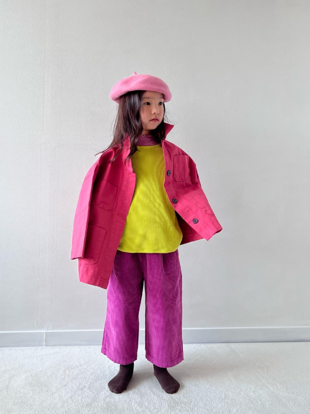 A child wearing wide-leg corduroy pants in magenta pink, paired with a bright yellow shirt, pink jacket and light pink beret. She is standing on grey carpet against a white wall..