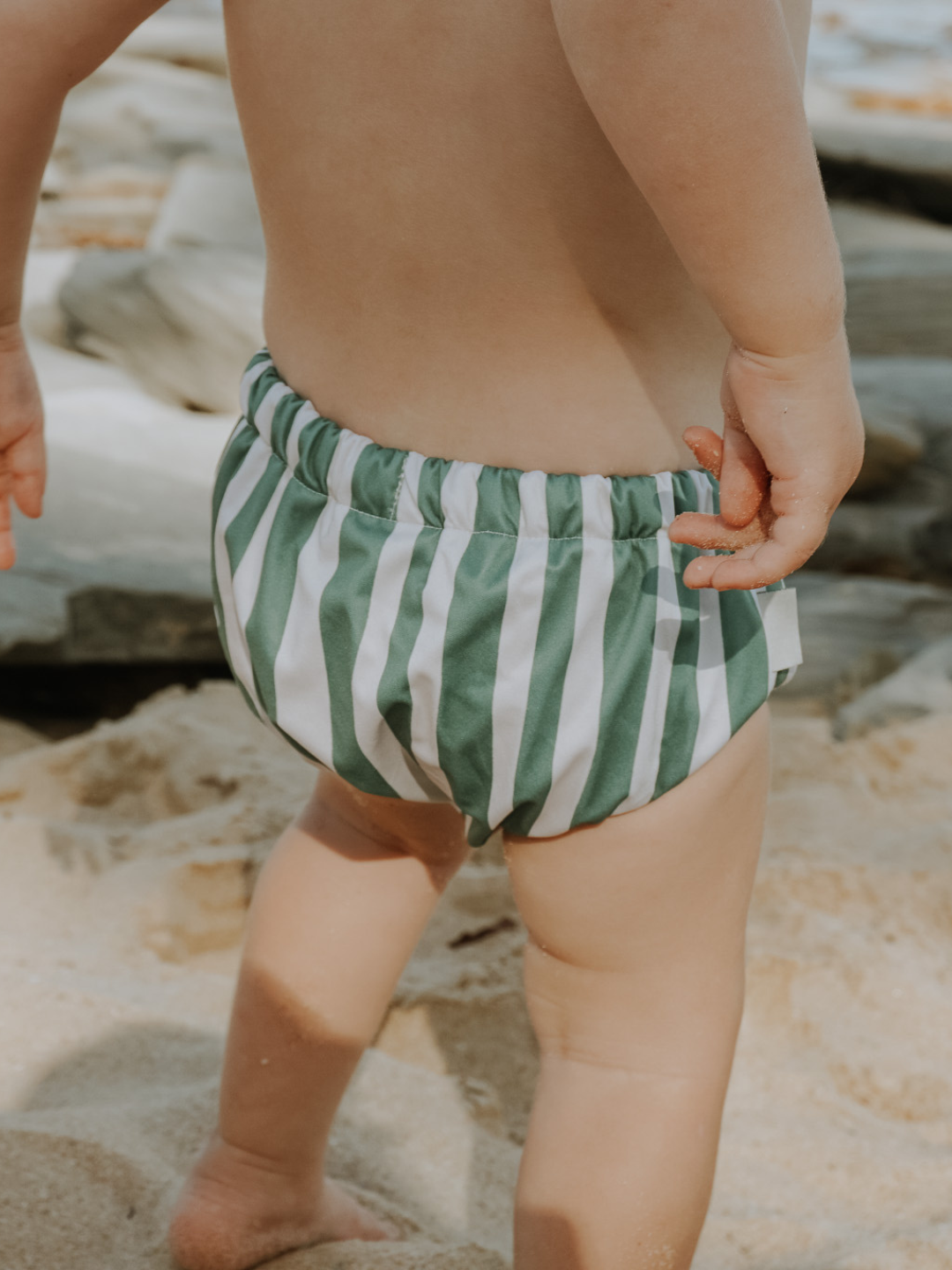The back view of the swim diaper with an elastic waist with a tie and elastic leg holes on a child at the beach.. The diaper is a has cream and moss green vertical stripes.