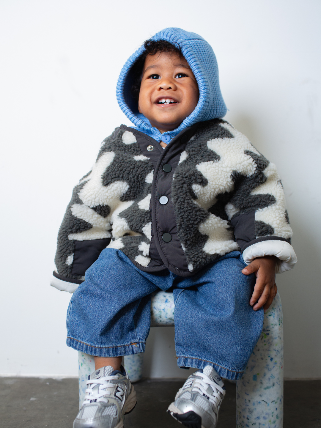 Child sitting on a small stool wearing a patterned jacket, sky blue hood and blue jeans against a plain background