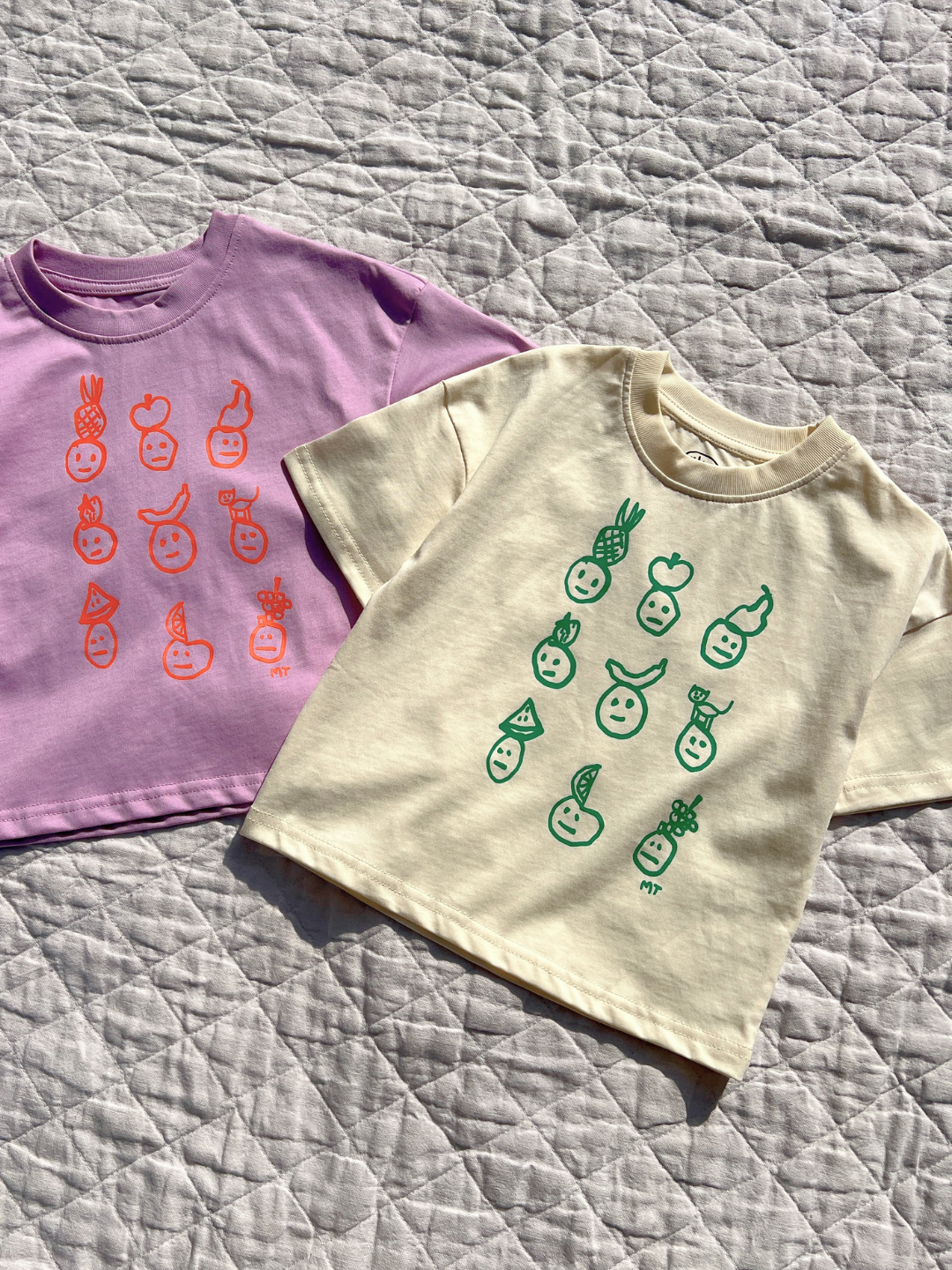 Both colorways of the Fruit Face tee are laid flat on the quilt in the sun.