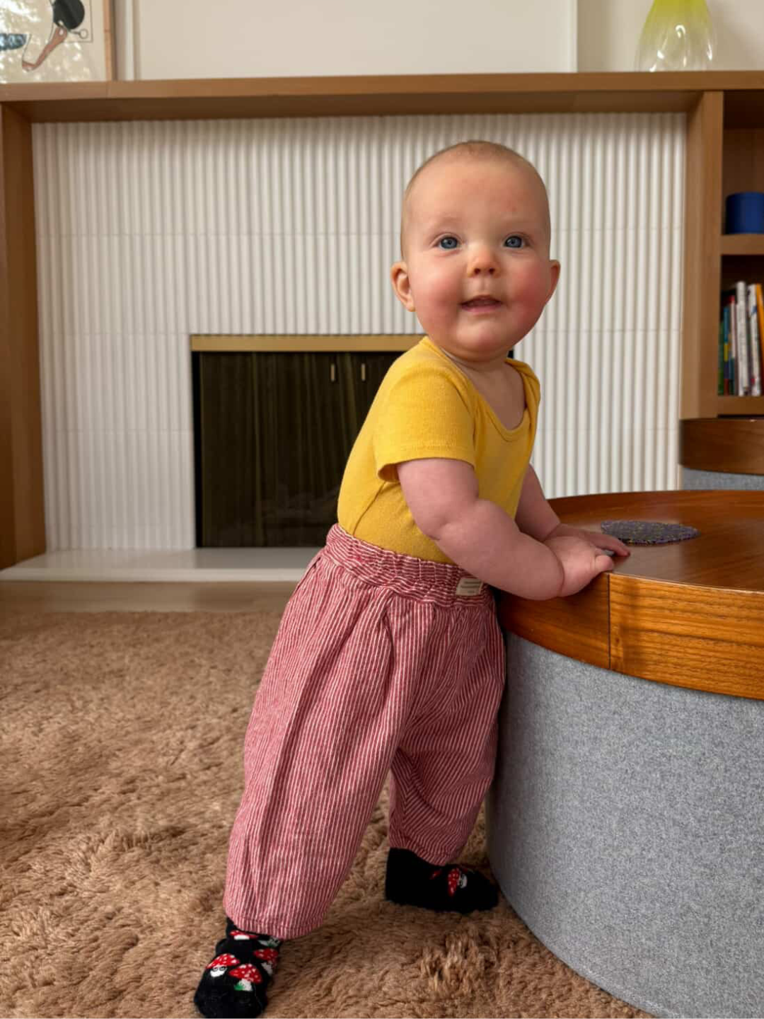 Baby in yellow shirt and red checkered pants standing next to a wooden desk.