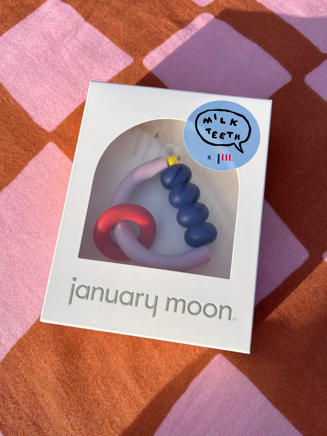A boxed MT X JM ARCH RING TEETHER, a US-made sensory-friendly teether, sits on pink and orange checkered fabric. The box features a “Milk Teeth” sticker and showcases the teether’s playful, colorful loops.