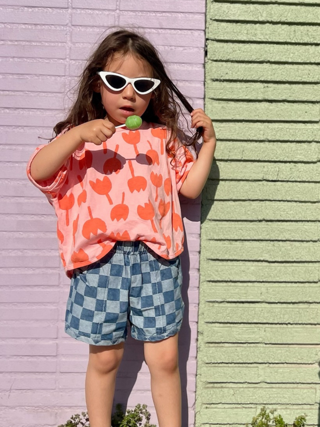 Child with long dark hair wearing the tulip tee with blue denim checkered shorts and white cat eye sunglasses. She is eating a green lollipop and standing in front of a purple and green painted brick wall.