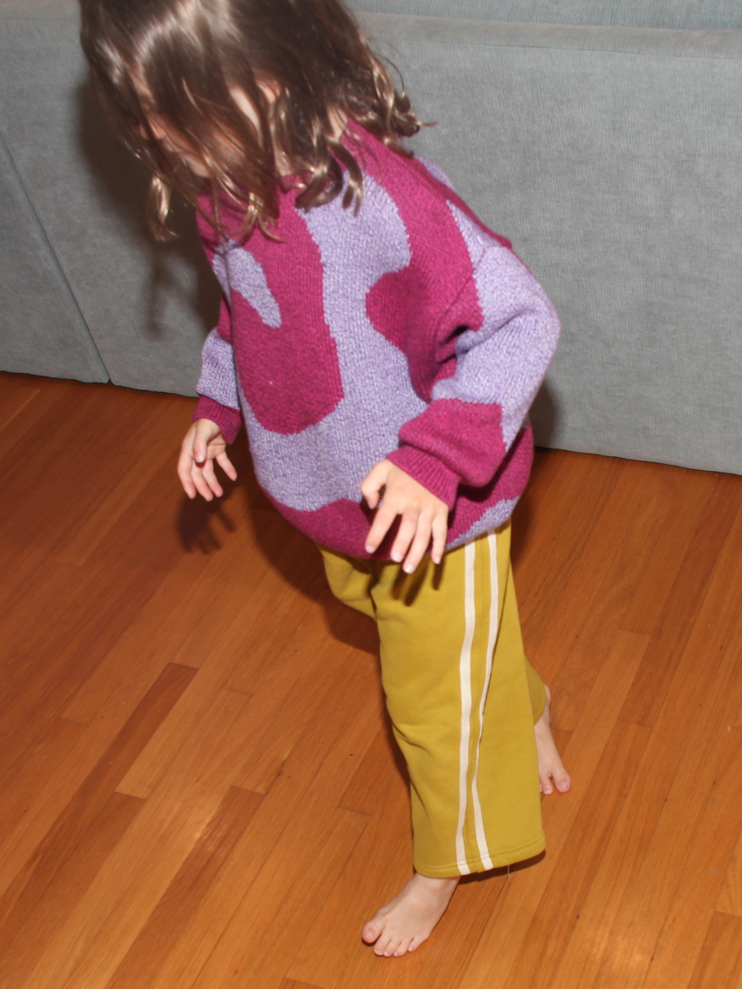 Chartreuse | A young child in SUNDAY TRACKPANTS and a purple and pink sweater walks barefoot on a wooden floor in front of a gray couch.