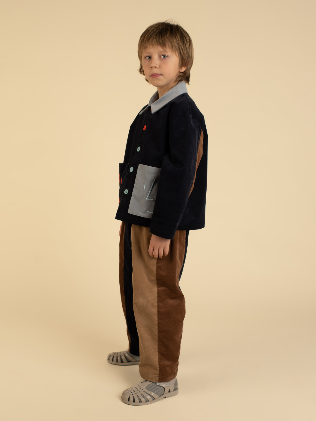 A child with light brown hair stands sideways, wearing the FACE JACKET and matching pants in navy, brown, and beige color-blocks, paired with light gray sandals, set against a beige background.