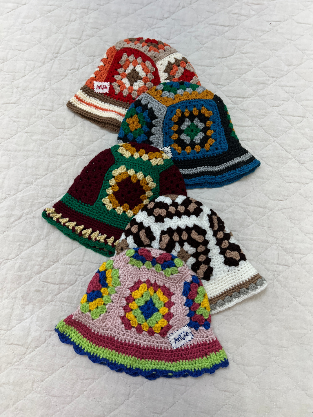 Four vibrant CROCHETED BUCKET HAT 1-3Y (Copy) hats with geometric South Africa-inspired patterns are arranged diagonally on a quilted white surface, highlighting their colorful, hand-made crochet design.
