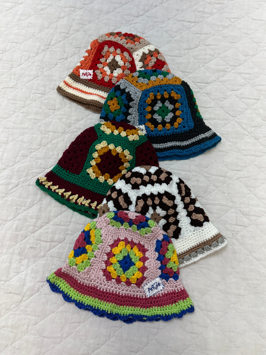 Image of Four vibrant CROCHETED BUCKET HAT 1-3Y (Copy) hats with geometric South Africa-inspired patterns are arranged diagonally on a quilted white surface, highlighting their colorful, hand-made crochet design.