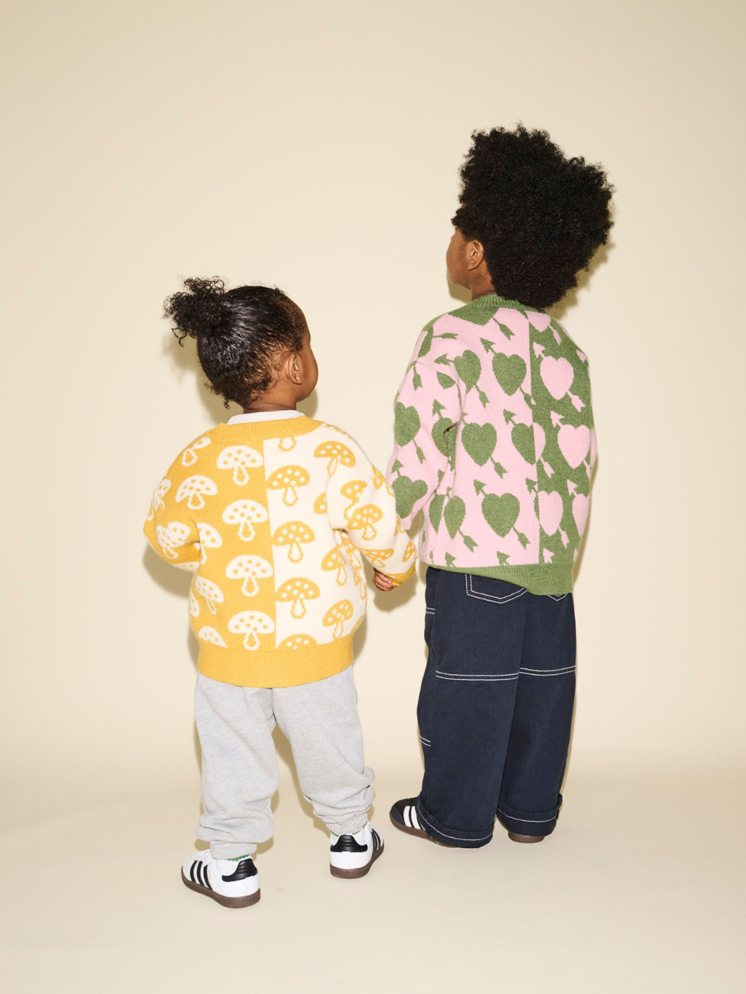 Two children standing and holding hands, facing away from the camera. One wears a SHROOM CARDIGAN featuring a charming yellow hue with a mushroom pattern, paired with gray pants and striped sneakers. The other child is dressed in a green sweater adorned with a pink heart pattern and dark blue pants—an adorable scene that needs no further description.