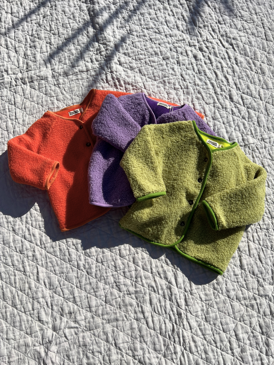 Matcha | Three kids collarless fleece jackets lying on an ecru quilt. The jackets are orange, purple and light green.