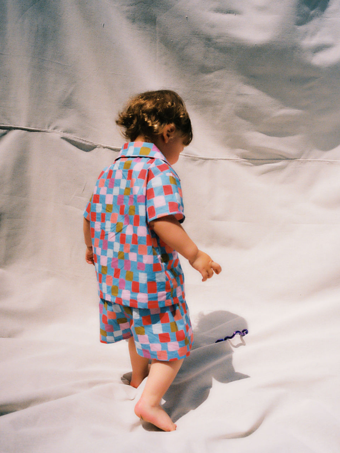 Blue | A toddler wearing a kids' shirt and shorts set in a pattern of red, orange, pink, lilac and green squares on a blue background, back view