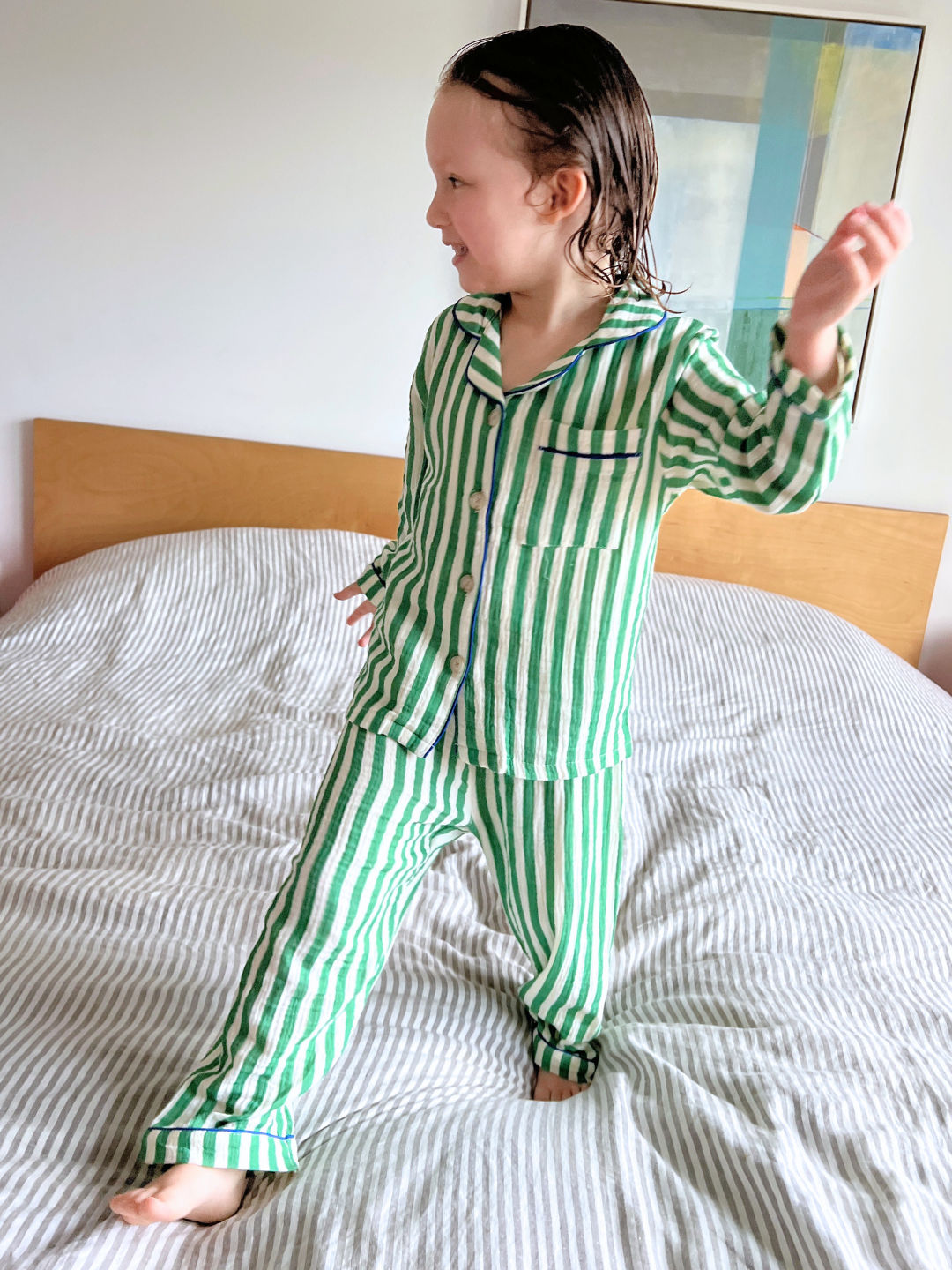 A child in a green and white striped Movie Night Set stands on a matching bed, with wet hair tousled, looking to the side and gesturing with one hand. The breathable fabric ensures comfort all night long.