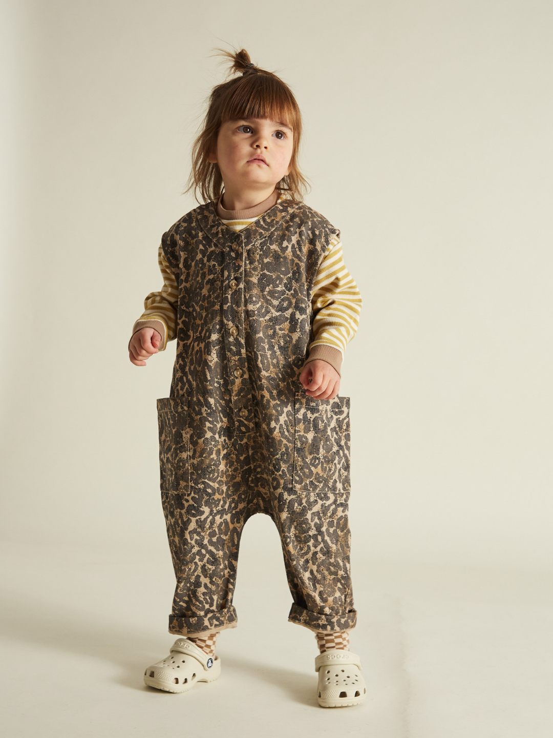 A toddler wears a Wren Leopard Oversized Overall by a unisex British brand, paired with a striped shirt and white clogs, styled with an adorable topknot.